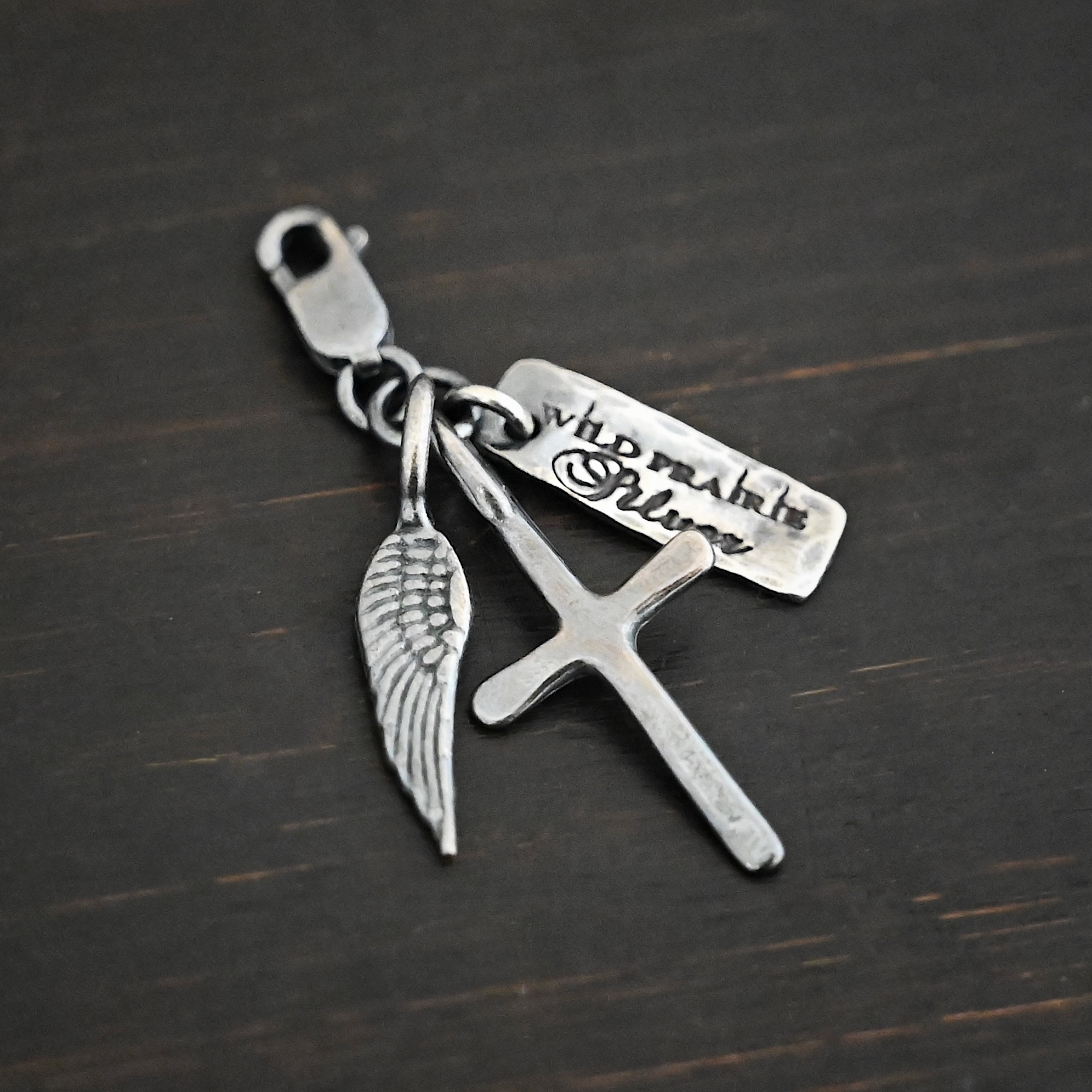 Wild Prairie Charm - oxidized shown on dark background - wild prairie tag, cross & angel wing charm