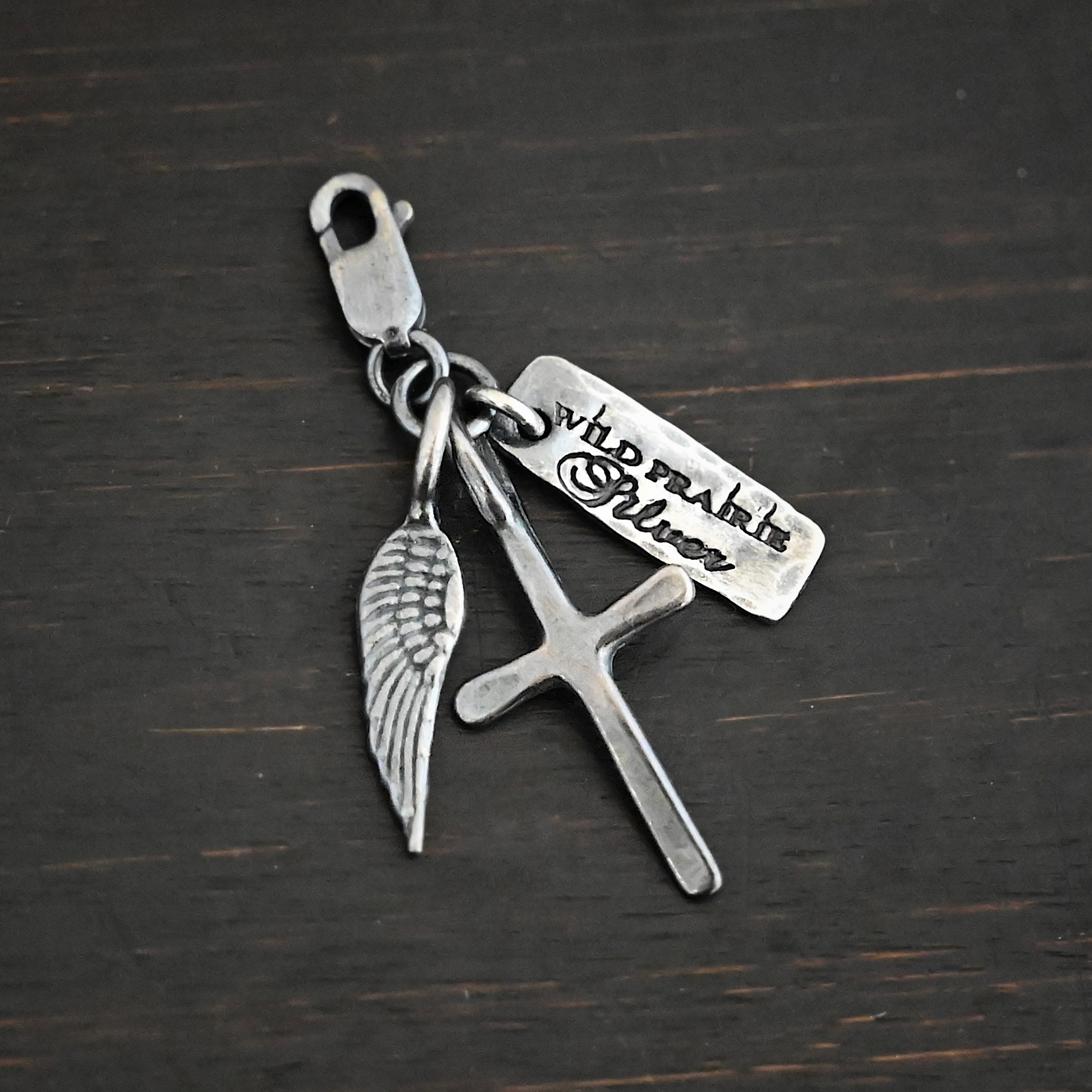 Wild Prairie Charm - oxidized shown on dark background - wild prairie tag, cross & angel wing charm