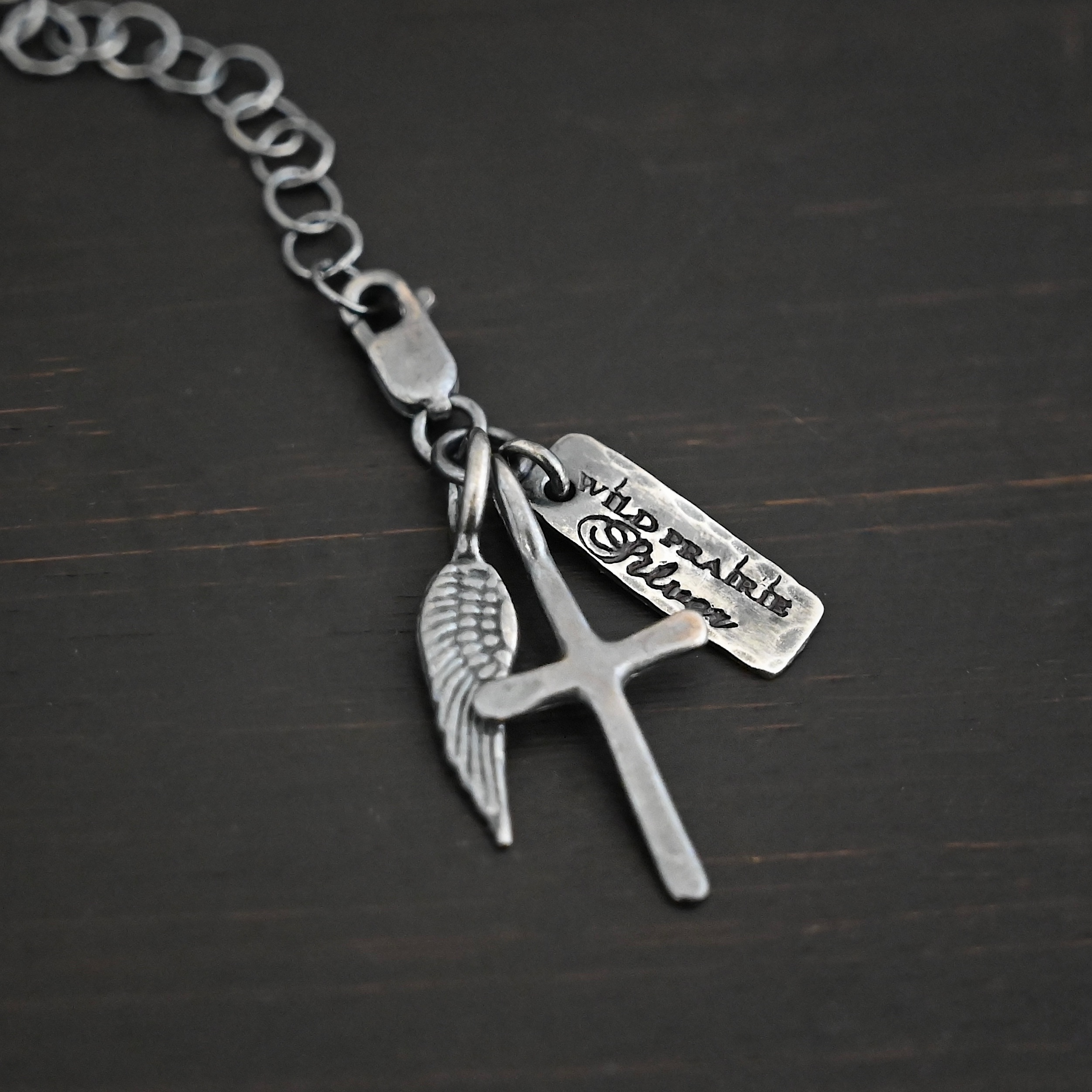 Wild Prairie Charm - oxidized shown on dark background - wild prairie tag, cross & angel wing charm