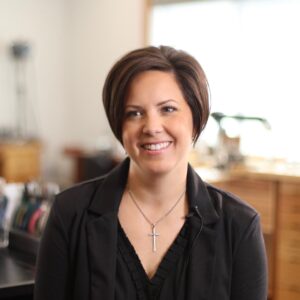 Kali Karnopp smiling at camera with short brown hair, cross necklace, and black jacket in Wild Prairie Studio
